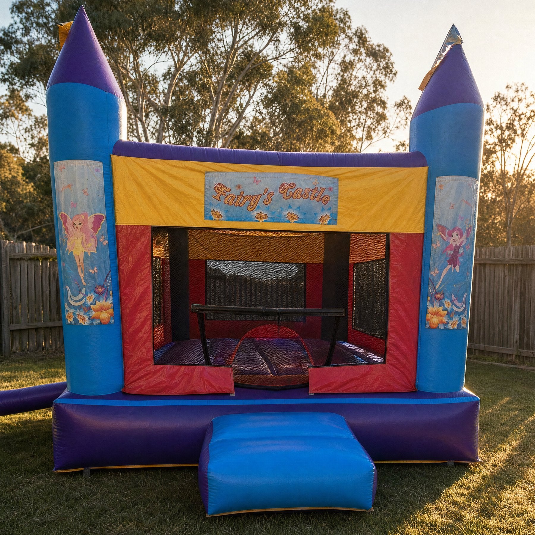 Purple and blue AM Banner jumping castle with Fairy's Castle banner in a backyard at golden hour