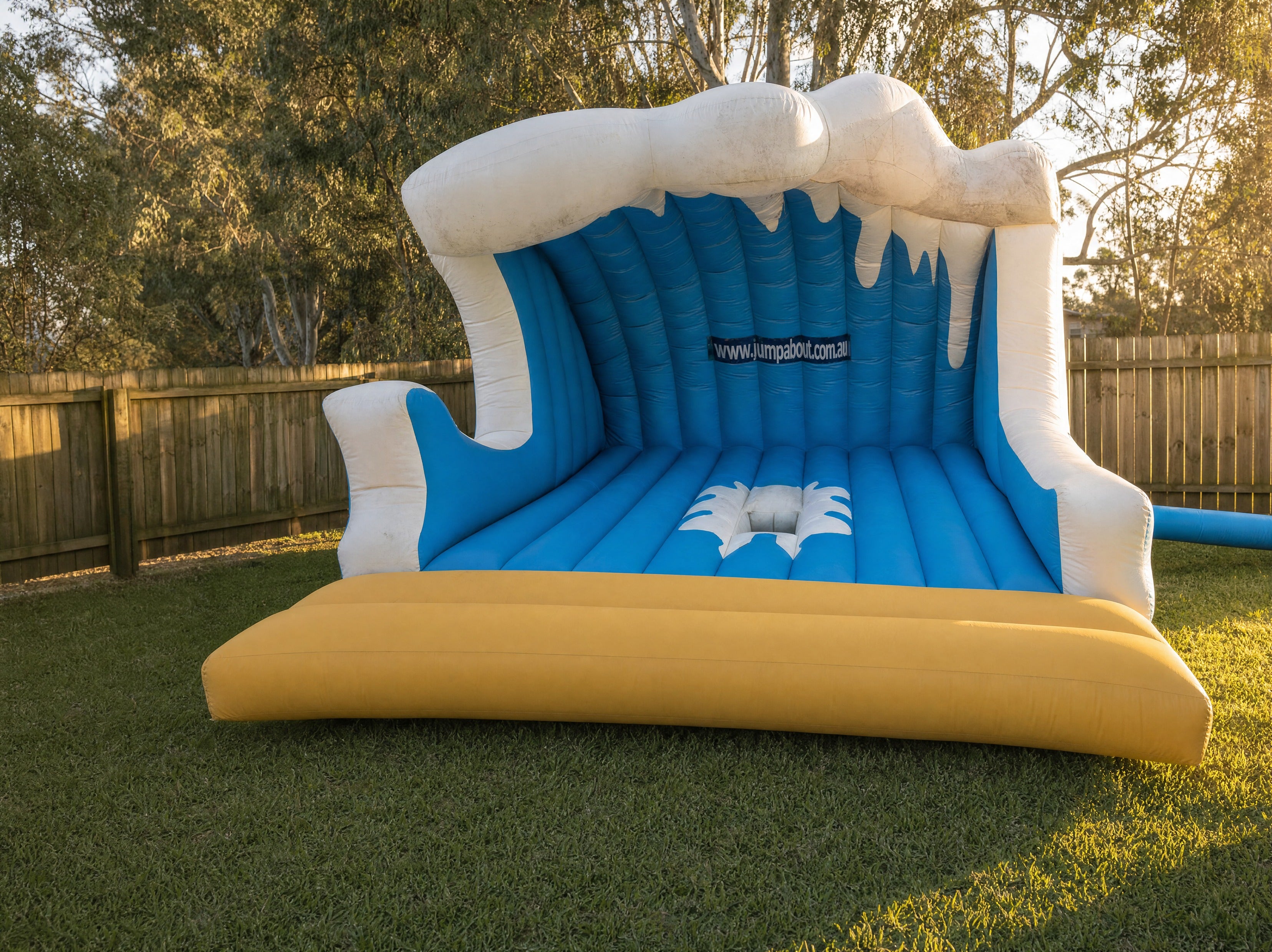 Mechanical Surfboard hire set up in a Central Coast backyard, front three-quarter view showing the full wave-shaped inflatable and landing mattress